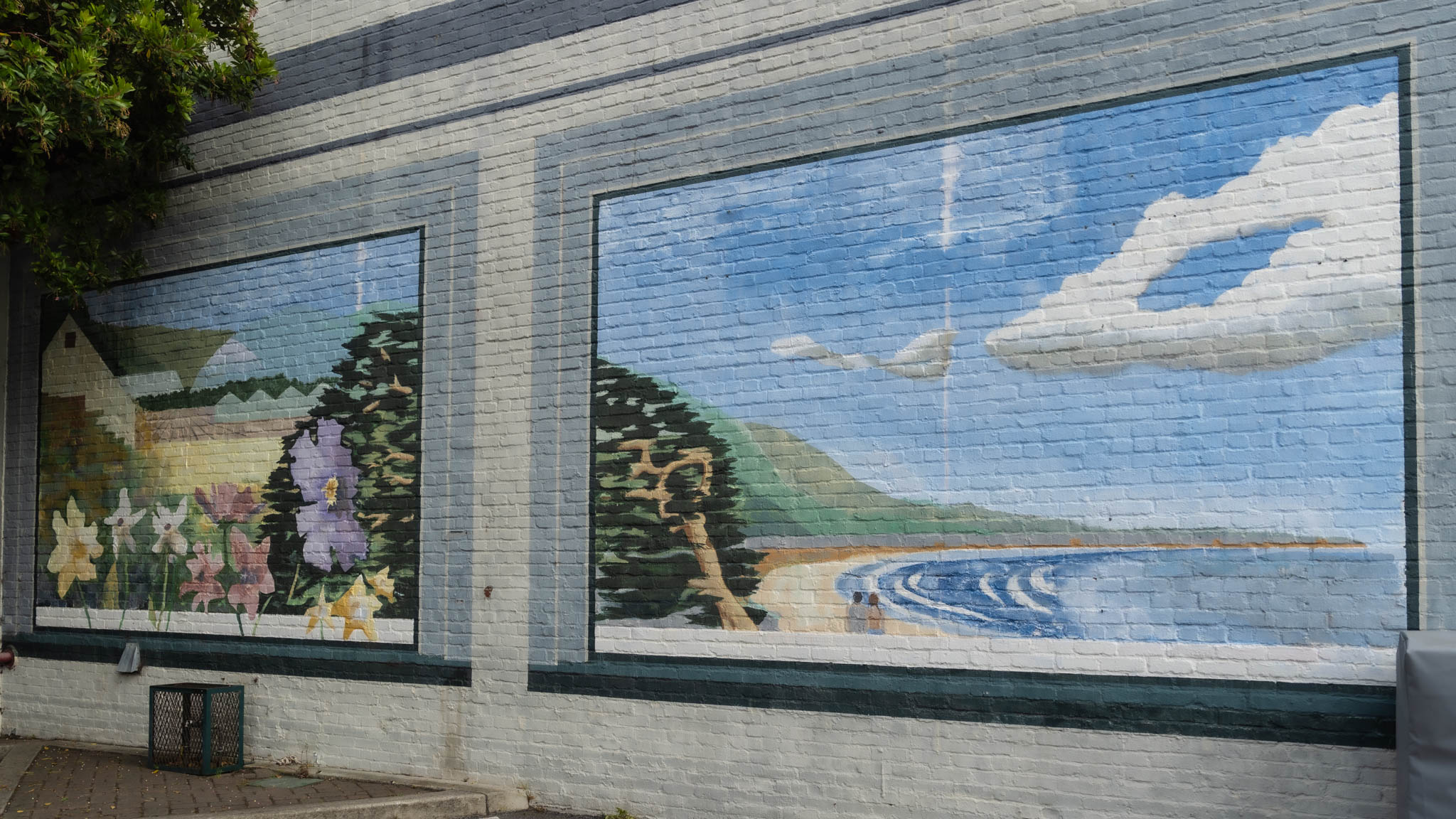 Mural on a brick wall showing flowers, trees, hills, a beach, and clouds in two framed scenes