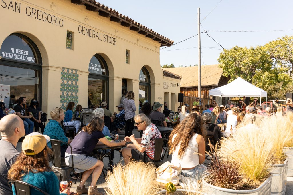 Outdoor gathering at San Gregorio General Store with people seated at tables and on chairs, facing a white canopy tent where a live band is performing. The building has a beige stucco exterior with arched windows and decorative tiles. Potted ornamental grasses line the foreground. The scene is sunny with clear skies.