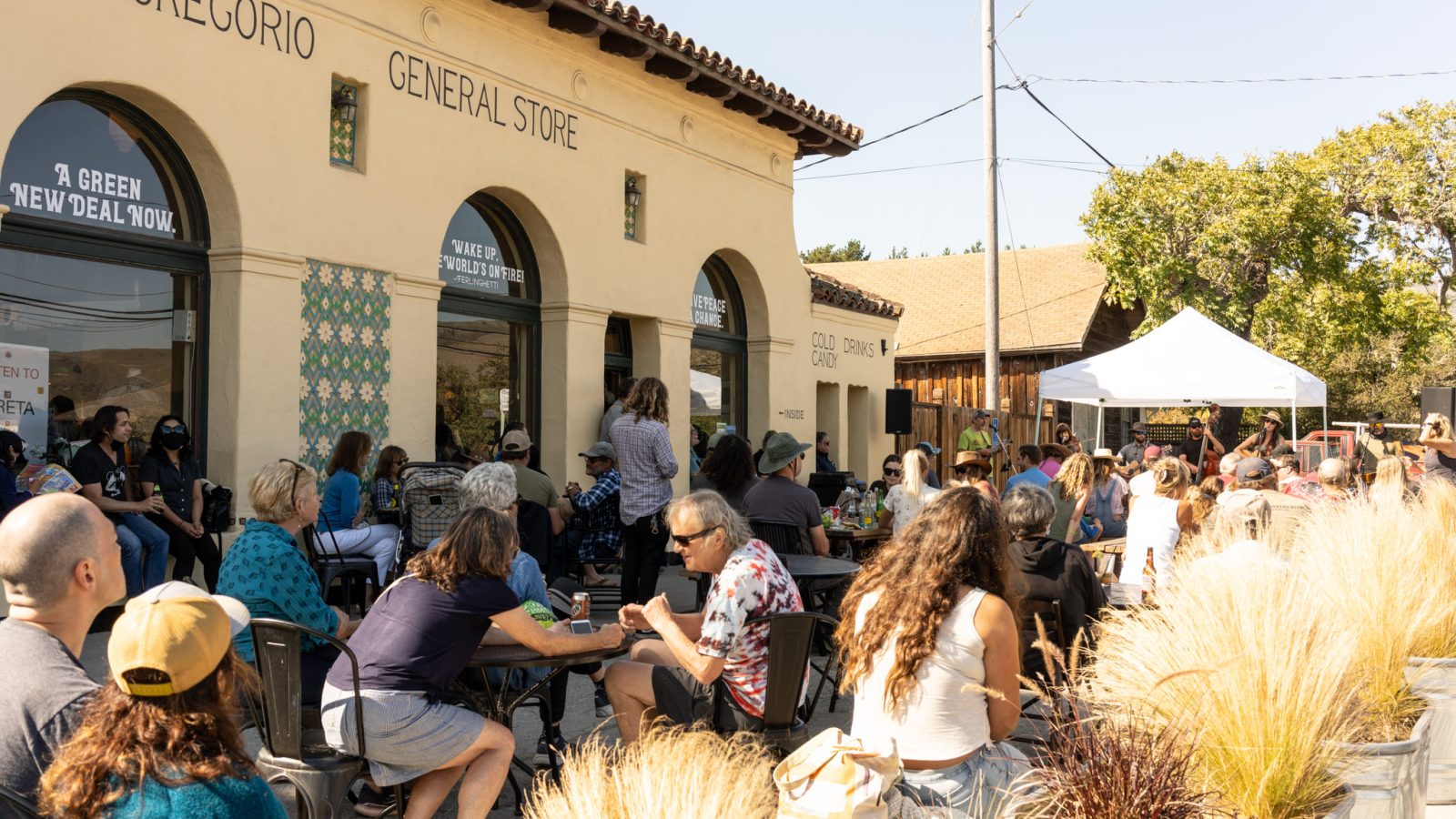 Outdoor gathering at San Gregorio General Store with people seated at tables and on chairs, facing a white canopy tent where a live band is performing. The building has a beige stucco exterior with arched windows and decorative tiles. Potted ornamental grasses line the foreground. The scene is sunny with clear skies.