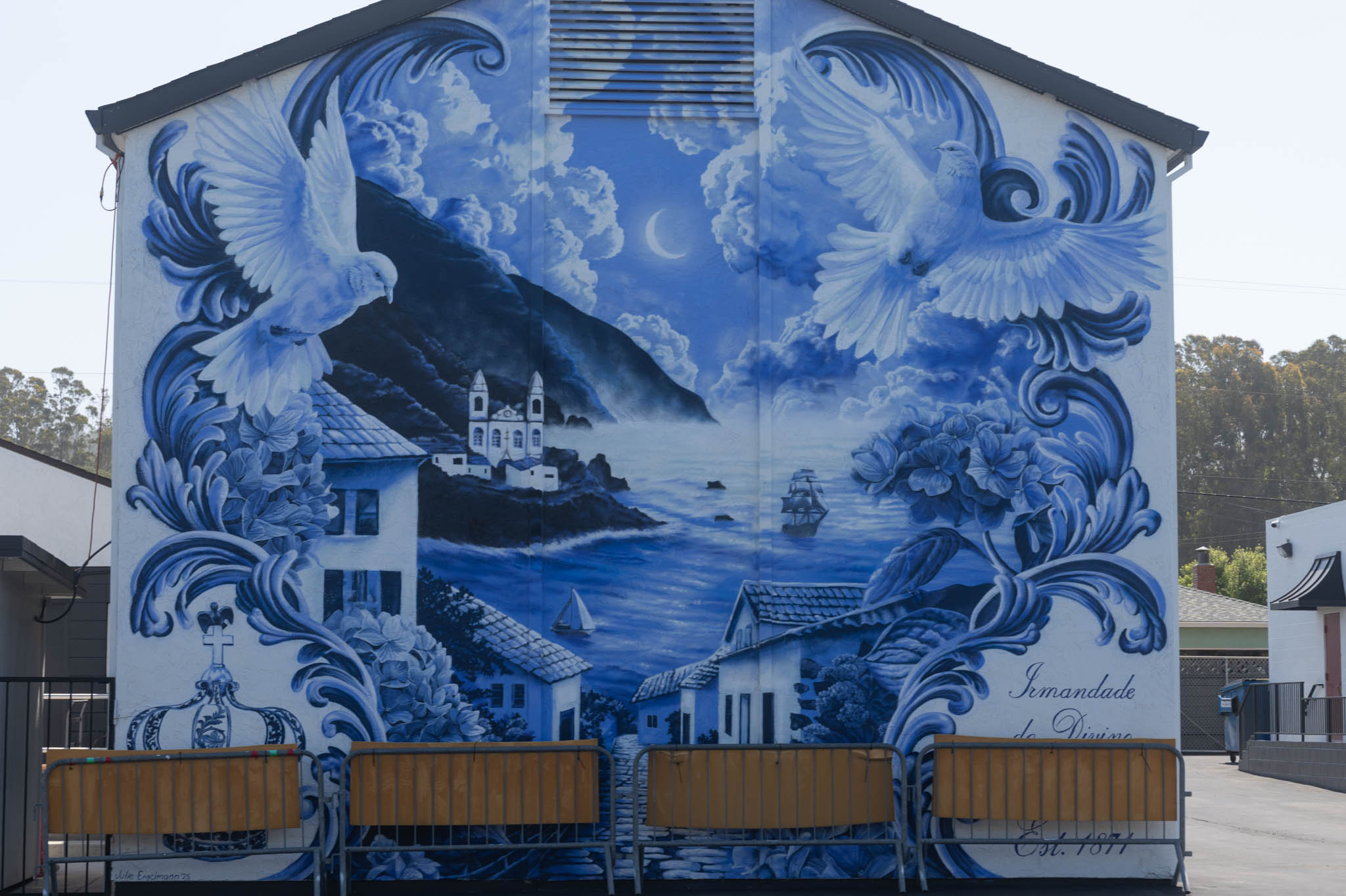 Blue mural on a building showing two white birds, a coastal village, mountains, clouds, and the text “Irmandade do Divino.”