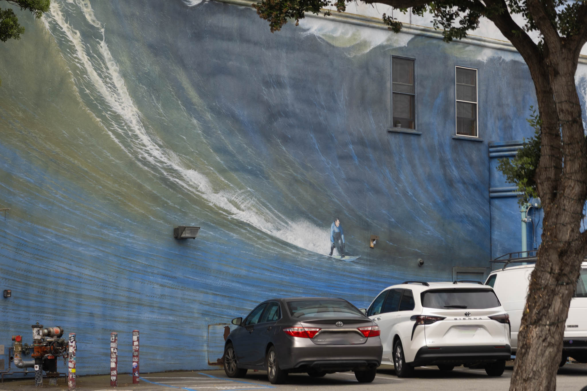 Large wave mural on a building wall behind parked cars and a tree, with a small surfer painted near the center.