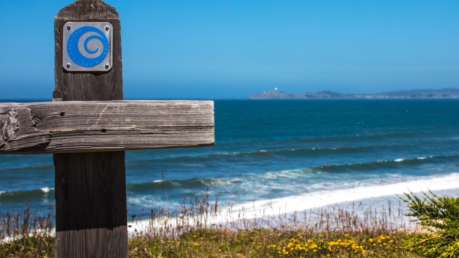Weathered wooden signpost with a blue circular California Coastal Trail emblem, set against a backdrop of ocean waves, a sandy beach with yellow wildflowers, and a clear blue sky.