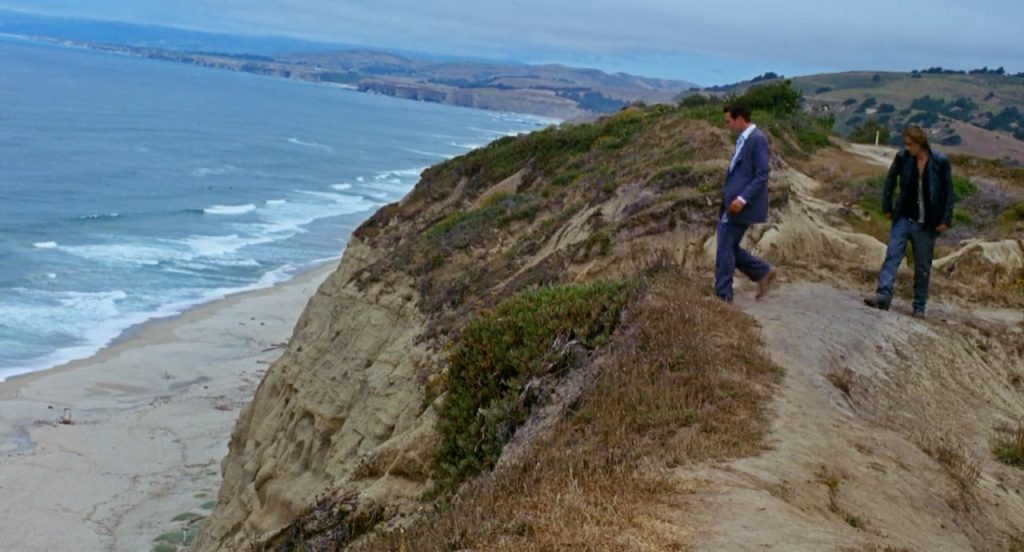 Two men walking on a dirt path along a grassy cliff overlooking a sandy beach and ocean waves, with hills visible in the background under a cloudy sky.