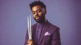 A man with short curly hair and a beard is wearing a purple suit jacket and matching shirt with a light purple pocket square. He is holding two white drumsticks vertically in front of him against a solid purple background.