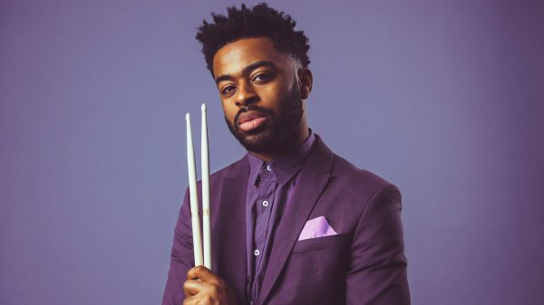 A man with short curly hair and a beard is wearing a purple suit jacket and matching shirt with a light purple pocket square. He is holding two white drumsticks vertically in front of him against a solid purple background.