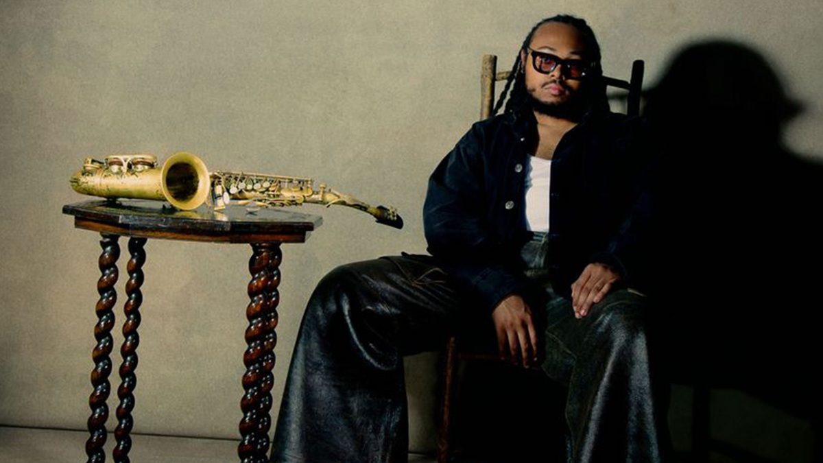 A man with braided hair and sunglasses sits on a wooden chair, wearing a dark jacket, white shirt, and shiny black pants. Next to him is a small wooden table with twisted legs, holding a brass saxophone. The background is plain and neutral-colored, casting a shadow behind the man.