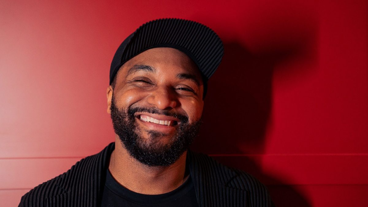 A smiling man with a beard wearing a black ribbed cap and a matching black ribbed jacket, standing against a red wall.