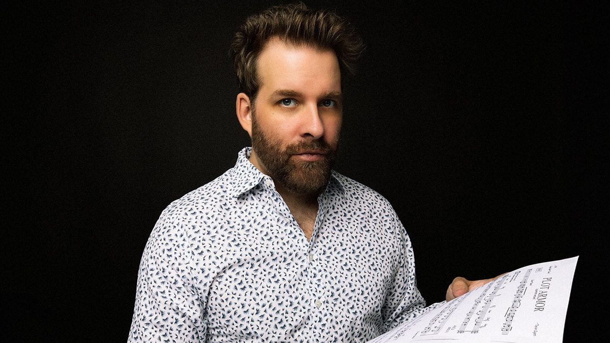 A man with a beard and styled hair is wearing a white shirt with a small dark pattern. He is holding a sheet of music titled "PLOT ARMOR" against a plain black background.