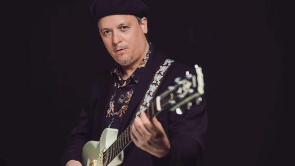 A man wearing a black beret and a dark blazer over a floral shirt is playing a light-colored electric guitar with a strap featuring a blue floral pattern. The background is solid black.