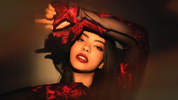 A woman with long dark hair wearing a sheer black top with red floral embroidery. She has bold red lipstick and is posing with one arm raised above her head, partially covering her forehead. The lighting casts warm shadows on her face and the background.