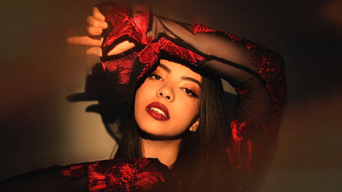 A woman with long dark hair wearing a sheer black top with red floral embroidery. She has bold red lipstick and is posing with one arm raised above her head, partially covering her forehead. The lighting casts warm shadows on her face and the background.
