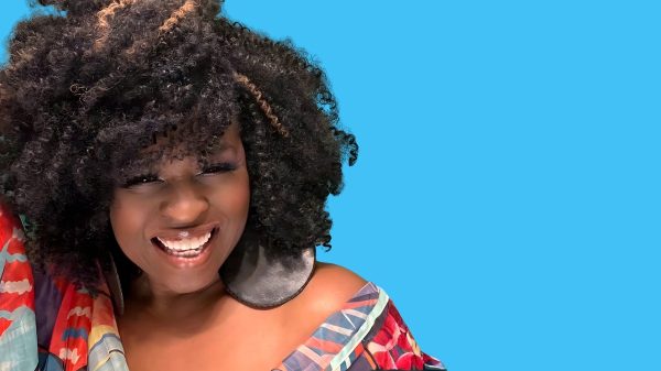 A smiling woman with voluminous curly hair featuring a few light brown highlights. She is wearing large round black earrings and a colorful off-the-shoulder top with a pattern of red, blue, and beige tones. The background is solid bright blue.