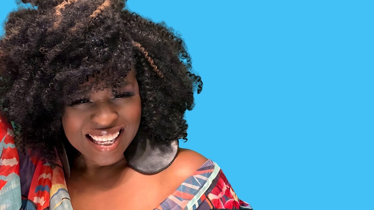 A smiling woman with voluminous curly hair featuring a few light brown highlights. She is wearing large round black earrings and a colorful off-the-shoulder top with a pattern of red, blue, and beige tones. The background is solid bright blue.