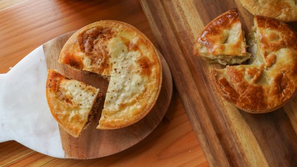 Two round meat pies with golden-brown crusts are placed on wooden boards. One pie on the left has a slice cut out and is on a board with a white marble handle, while the other pie on the right has a slice removed and is on a rectangular wooden board. The pies have a flaky, slightly browned top crust.