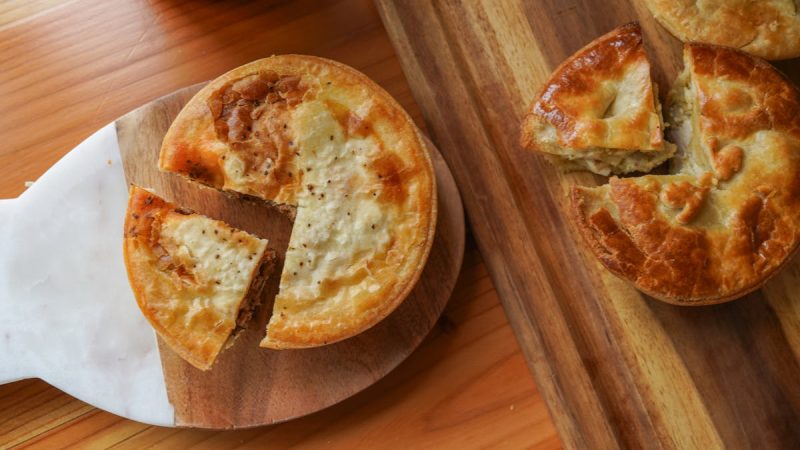 Two round meat pies with golden-brown crusts are placed on wooden boards. One pie on the left has a slice cut out and is on a board with a white marble handle, while the other pie on the right has a slice removed and is on a rectangular wooden board. The pies have a flaky, slightly browned top crust.