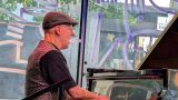 Man in a flat cap plays a Yamaha grand piano by a window with purple painted designs.