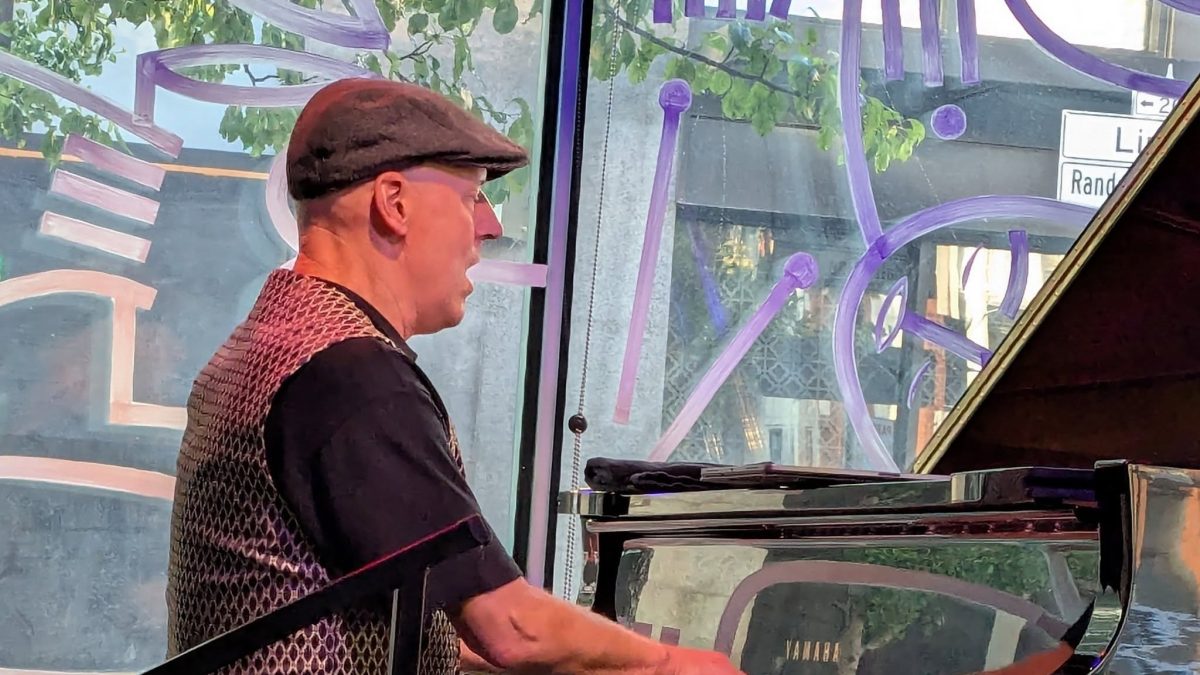 Man in a flat cap plays a Yamaha grand piano by a window with purple painted designs.