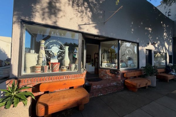 A ceramics gallery storefront with large display windows showcasing various pottery pieces, including vases and pots. The building has a light-colored exterior with brick accents below the windows. There are three wooden benches outside along the sidewalk, and a potted plant is placed near the left bench. The gallery entrance is open, revealing more pottery inside. The windows have circular logos with text indicating "Jesse Henry Ceramics Miwok Gallery Half Moon Bay, CA" and "Kamal Reid Ceramics Studio.