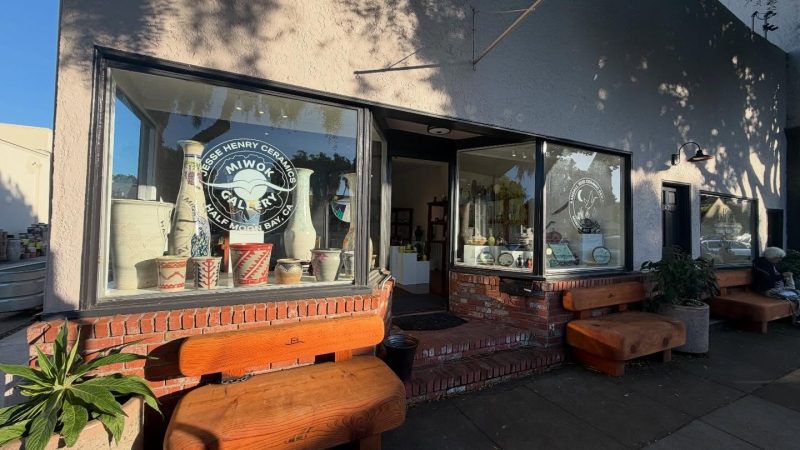A ceramics gallery storefront with large display windows showcasing various pottery pieces, including vases and pots. The building has a light-colored exterior with brick accents below the windows. There are three wooden benches outside along the sidewalk, and a potted plant is placed near the left bench. The gallery entrance is open, revealing more pottery inside. The windows have circular logos with text indicating "Jesse Henry Ceramics Miwok Gallery Half Moon Bay, CA" and "Kamal Reid Ceramics Studio.