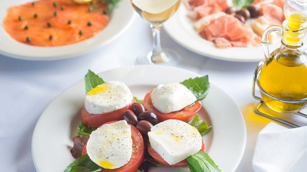 A plate with four slices of tomato topped with fresh mozzarella cheese, basil leaves, and a drizzle of olive oil, accompanied by black olives in the center. In the background, there is a glass of white wine, a plate of smoked salmon garnished with capers and a lemon wedge, and another plate with slices of prosciutto and olives. A bottle of olive oil is also visible on the right side.