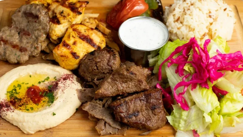 Grilled mixed meat platter with chicken, beef, and lamb pieces, served with hummus topped with olive oil and spices, a side of rice, grilled tomato and pepper, a small cup of white sauce, and a fresh salad with lettuce and shredded purple cabbage on a wooden board.