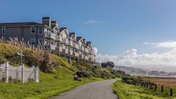 A row of beige multi-story townhouses with balconies sits atop a grassy hill beside a paved pathway. The scene is set under a clear blue sky with some clouds in the distance, and there is a white picket fence and tall ornamental grasses near the houses. The pathway curves gently and is bordered by green grass on both sides.