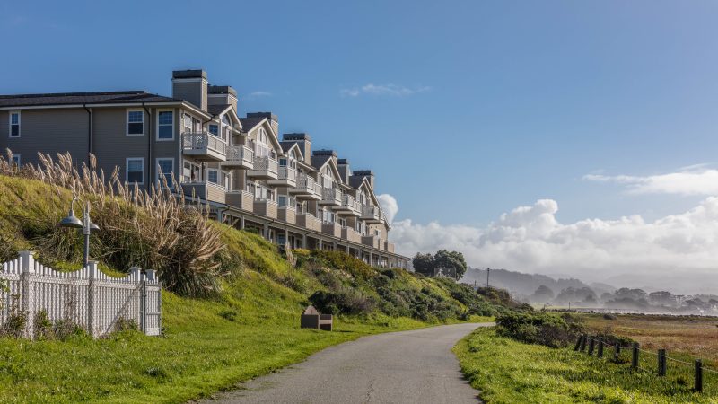 A row of beige multi-story townhouses with balconies sits atop a grassy hill beside a paved pathway. The scene is set under a clear blue sky with some clouds in the distance, and there is a white picket fence and tall ornamental grasses near the houses. The pathway curves gently and is bordered by green grass on both sides.