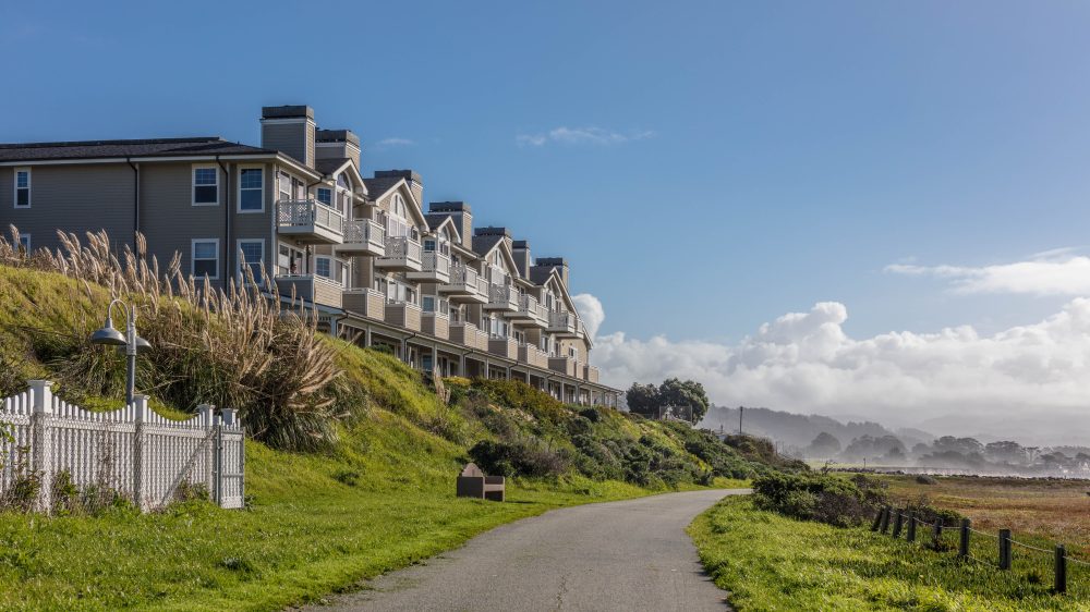 A row of beige multi-story townhouses with balconies sits atop a grassy hill beside a paved pathway. The scene is set under a clear blue sky with some clouds in the distance, and there is a white picket fence and tall ornamental grasses near the houses. The pathway curves gently and is bordered by green grass on both sides.
