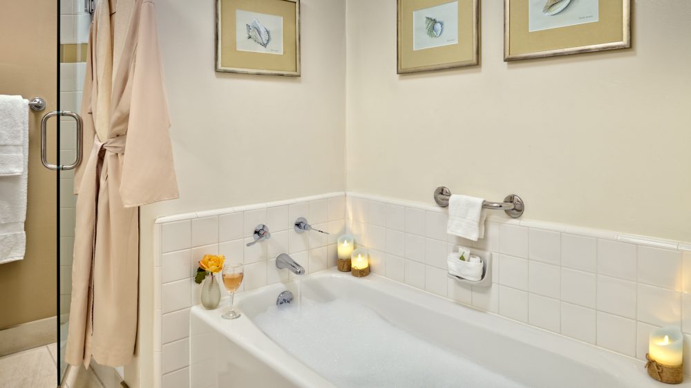 Bathtub filled with bubbles and running water, surrounded by white tiled walls. A beige bathrobe hangs on a hook near a glass shower door. Three framed pictures of seashells are mounted on the wall above the tub. Two lit candles and a small vase with a yellow flower are placed on the edge of the tub, along with a glass of wine. A towel rack with a white towel and a soap holder with a small towel are mounted on the wall.