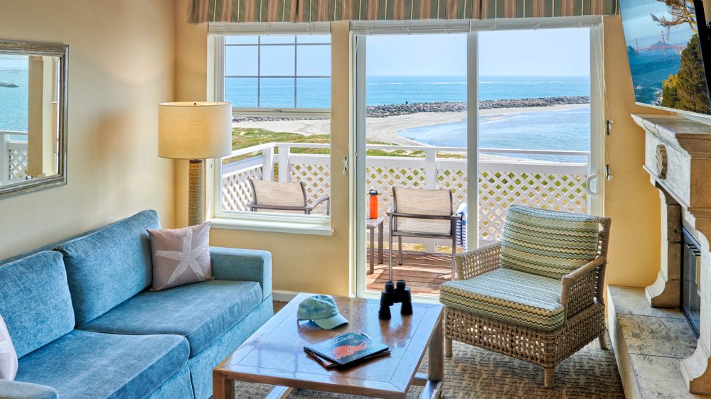 Cozy living room with a blue sofa, a wicker armchair with patterned cushions, and a wooden coffee table holding a blue cap, binoculars, and a book. A floor lamp stands next to the sofa, and a large window and sliding glass door offer a view of a balcony with two chairs and a table, overlooking a sandy beach and calm ocean. A flat-screen TV is mounted above a stone fireplace.
