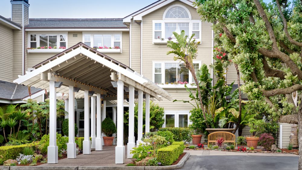 A beige multi-story building with white-framed windows and flower boxes beneath them. In front, there is a white covered walkway supported by multiple square columns. The area around the entrance is landscaped with green shrubs, potted plants, and trees, including tall tropical plants and a large tree with red flowers. A wooden bench is placed near the plants on the right side. The sky is clear and blue.
