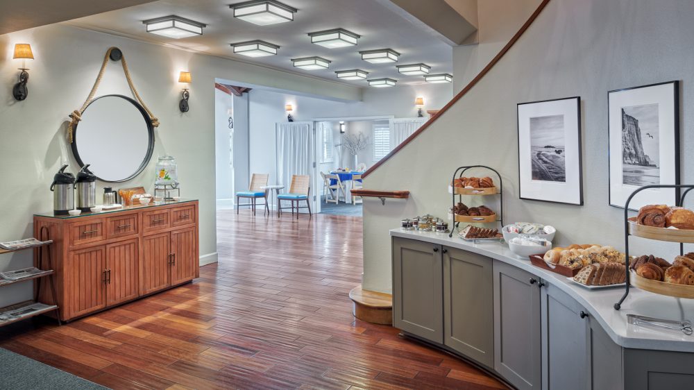 A breakfast buffet area with wooden flooring and soft lighting. On the right, a curved gray countertop holds various pastries, bread, and spreads, displayed on tiered trays and plates. Above the counter are two black-and-white framed photographs of coastal scenes. On the left, a wooden cabinet with a round mirror hanging above it has coffee dispensers, a glass water dispenser with lemon slices, and small bowls with condiments. In the background, there are two chairs with blue cushions and a table set for dining in a bright room.