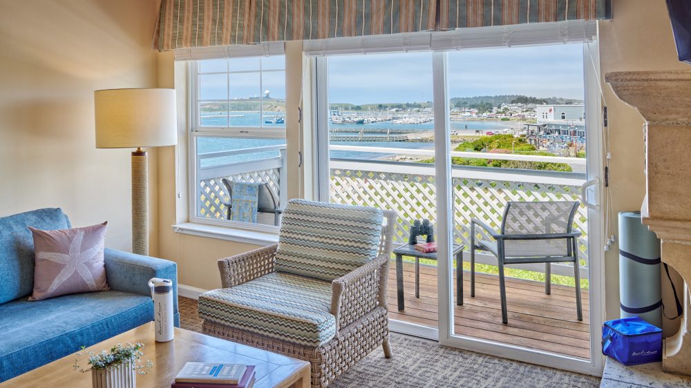 Living room with a blue sofa featuring a starfish pillow, a wicker armchair with patterned cushions, a wooden coffee table with books and a small plant, and a tall floor lamp. Sliding glass doors and a window offer a view of a balcony with outdoor chairs and a table, overlooking a marina and waterfront area. The room has beige walls and a patterned carpet.