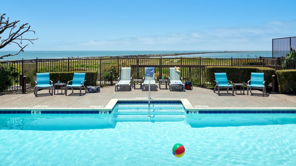 Outdoor swimming pool with clear blue water and a colorful beach ball floating on the surface. Surrounding the pool are several lounge chairs, some with towels and a hat, arranged on a concrete deck. Beyond the pool area is a black metal fence, green grass, and a view of the ocean under a clear blue sky.