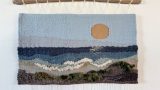 Woven wall hanging depicting a landscape scene with a large orange sun in a light blue sky, layered blue and gray waves, white foamy surf, and a dark brown and green textured shoreline. The tapestry is suspended from a natural wooden branch with cream-colored hanging cords.