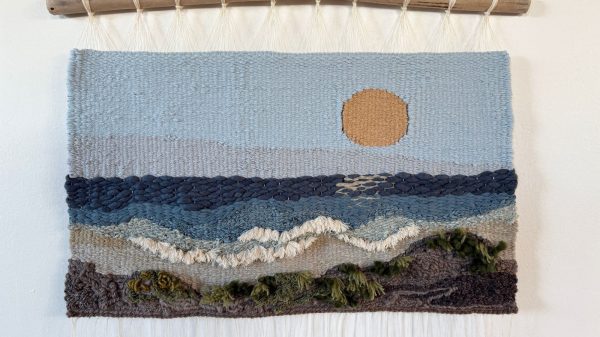 Woven wall hanging depicting a landscape scene with a large orange sun in a light blue sky, layered blue and gray waves, white foamy surf, and a dark brown and green textured shoreline. The tapestry is suspended from a natural wooden branch with cream-colored hanging cords.