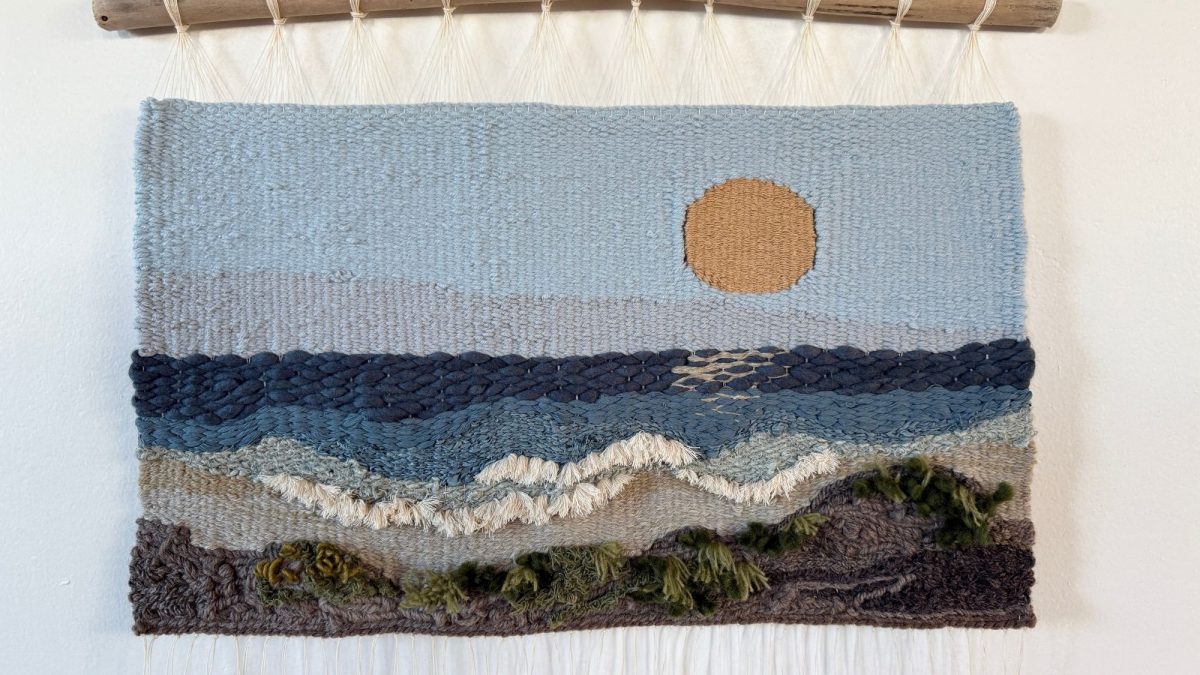 Woven wall hanging depicting a landscape scene with a large orange sun in a light blue sky, layered blue and gray waves, white foamy surf, and a dark brown and green textured shoreline. The tapestry is suspended from a natural wooden branch with cream-colored hanging cords.