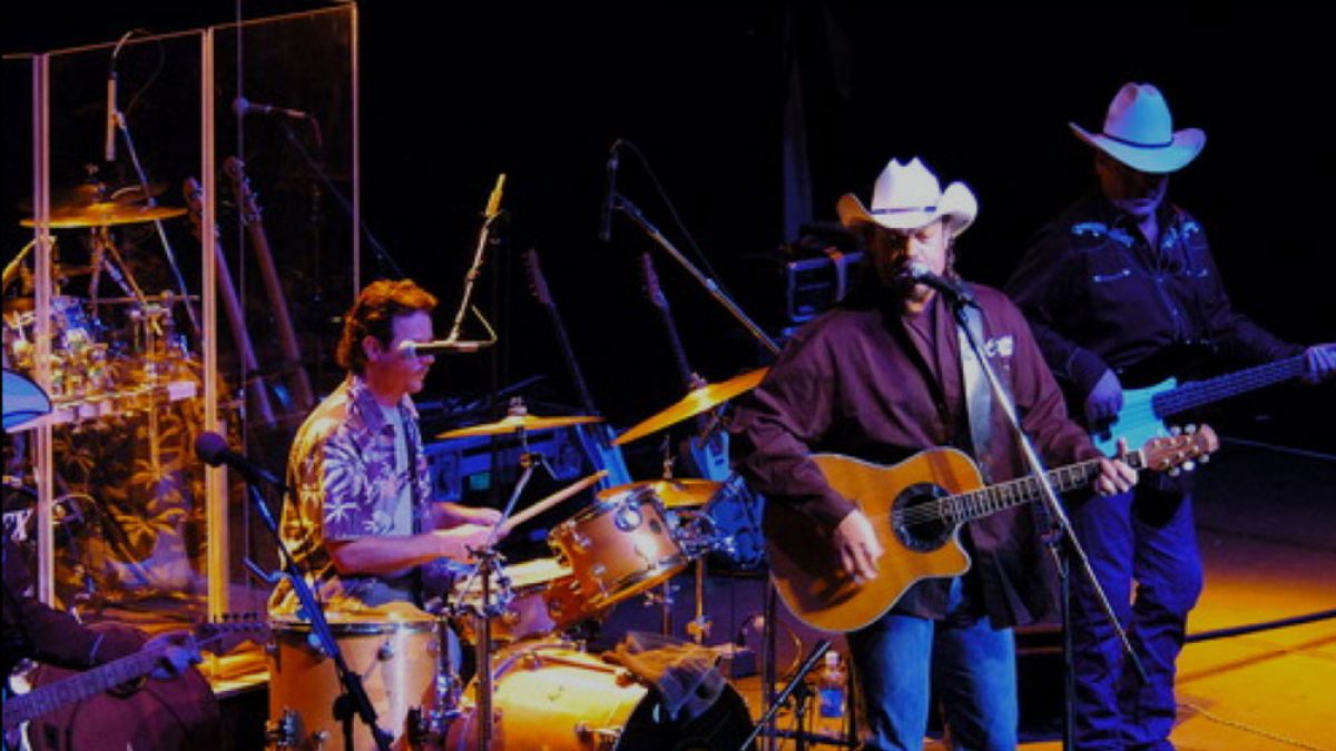 A country band performing on stage, featuring a drummer in a patterned shirt, a guitarist and singer wearing a white cowboy hat and dark shirt, and a bassist also wearing a white cowboy hat and dark clothing. The stage is dimly lit with warm lighting focused on the musicians.