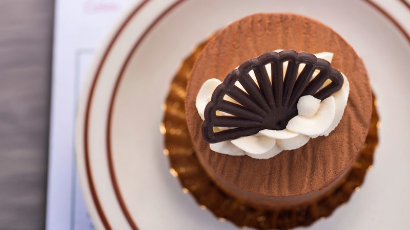 A round chocolate mousse cake topped with white cream swirls and a decorative dark chocolate piece shaped like a fan, placed on a gold foil base and a white plate with brown rings.