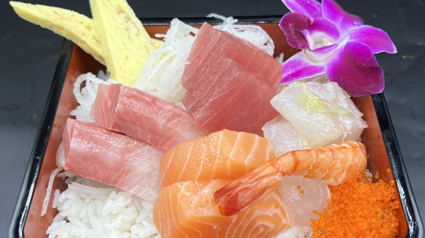 A black rectangular container filled with sushi rice topped with various sashimi pieces including salmon, tuna, shrimp, and white fish. There are two slices of tamago (Japanese omelette), a small mound of orange fish roe, shredded daikon radish, and a purple orchid flower for garnish. The background is black.