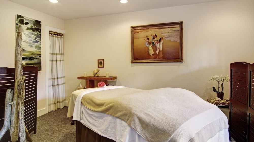 A cozy massage room featuring a massage table covered with white and beige linens, a small wooden side table with candles and a flower, and a potted orchid on a stool. The walls are light-colored with a framed painting of people on a beach and another landscape painting. The room is softly lit by recessed ceiling lights and has wooden folding screens on either side.