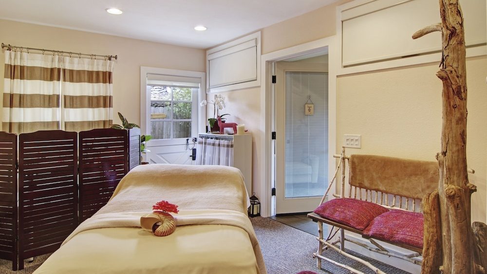 Massage room with a massage table covered in beige sheets and a beige blanket, topped with a seashell and a red flower. A wooden folding screen with horizontal slats is positioned near a window with striped beige and white curtains. A rustic wooden bench with red cushions and a brown throw is against the wall, next to a tall, natural wooden coat rack. The room has beige walls, recessed ceiling lights, and a glass door leading to another area.