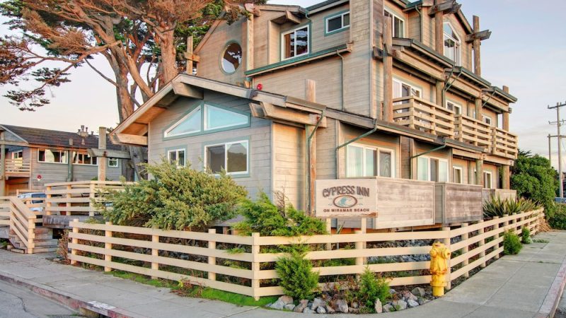 A multi-story wooden building with beige siding and green trim, surrounded by a light wooden fence and greenery. The building has several windows, balconies, and a sign that reads "Cypress Inn on Miramar Beach." There is a yellow fire hydrant near the corner of the fence, and large trees are visible behind the building. The scene is set on a paved sidewalk and street.