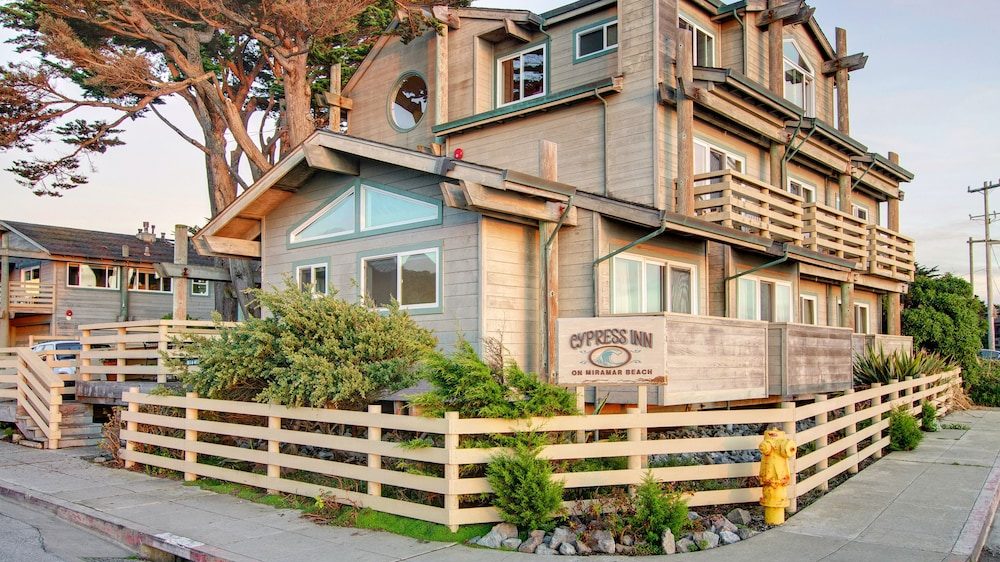 A multi-story wooden building with beige siding and green trim, surrounded by a light wooden fence and greenery. The building has several windows, balconies, and a sign that reads "Cypress Inn on Miramar Beach." There is a yellow fire hydrant near the corner of the fence, and large trees are visible behind the building. The scene is set on a paved sidewalk and street.