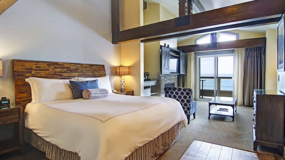 Bedroom with a large wooden headboard bed dressed in white linens and decorative pillows, flanked by wooden nightstands with lamps. The room features exposed wooden beams, a patterned armchair, a glass coffee table, a dresser, and sliding glass doors leading to a balcony with an ocean view.