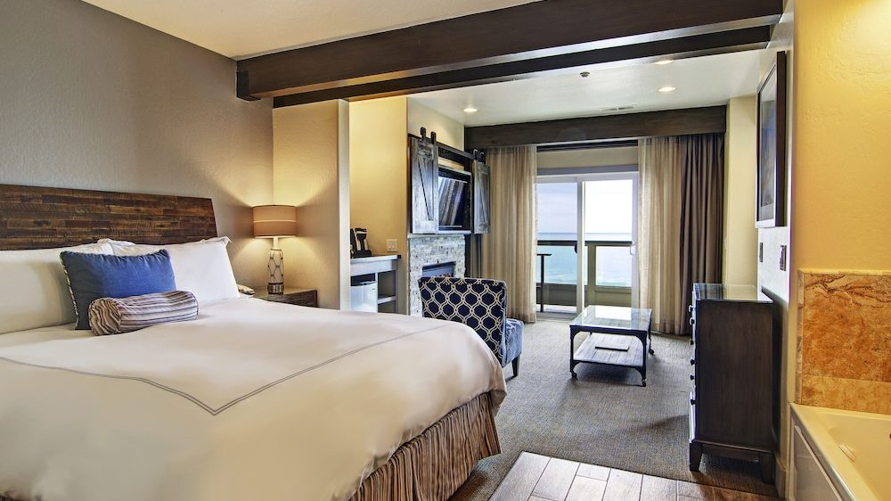 Hotel room with a large bed featuring white linens and decorative pillows, a wooden headboard, a bedside lamp on a small table, a patterned armchair, a coffee table, a dresser, and a wall-mounted TV with sliding barn-style doors. The room has carpeted flooring near the seating area and wood-like flooring near the entrance, with a sliding glass door leading to a balcony overlooking the ocean.