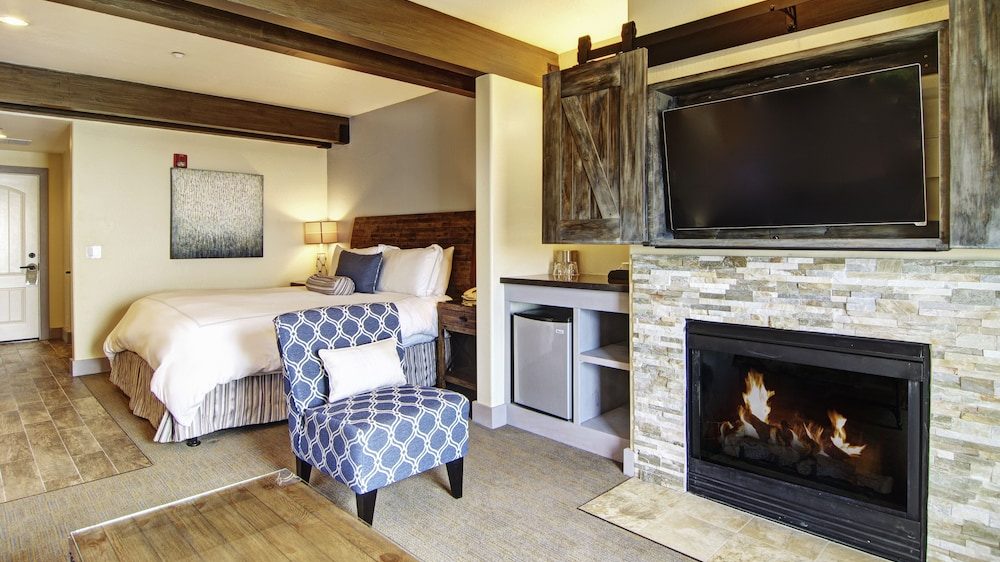 A cozy hotel room featuring a neatly made bed with white linens and blue accent pillows, a blue patterned armchair with a white cushion, a wooden coffee table, and a stone fireplace with a fire burning. Above the fireplace is a flat-screen TV mounted inside a rustic wooden cabinet with sliding barn-style doors. The room has wooden ceiling beams, a small refrigerator, and soft lighting from a bedside lamp.