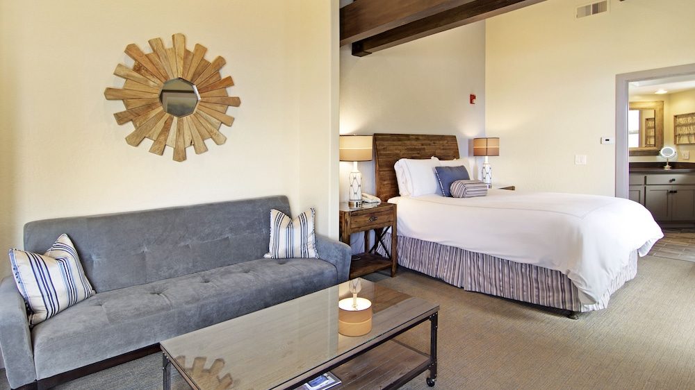 A bedroom with a wooden headboard bed dressed in white linens and striped bed skirt, flanked by two wooden nightstands each with a lamp. In front of the bed is a gray upholstered sofa with striped pillows. A glass-top coffee table with a candle sits in front of the sofa. A decorative wooden sunburst mirror hangs on the wall above the sofa. The room has beige walls and carpet, with an open doorway leading to a bathroom.