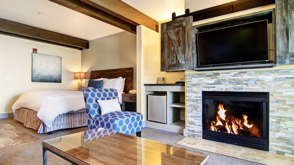 A cozy bedroom featuring a large bed with white bedding and a wooden headboard, a blue patterned armchair with a white pillow, a glass coffee table, and a stone fireplace with a fire burning. Above the fireplace is a flat-screen TV mounted inside rustic wooden sliding barn doors. The room has exposed wooden ceiling beams and a small kitchenette area with a mini fridge and shelves.
