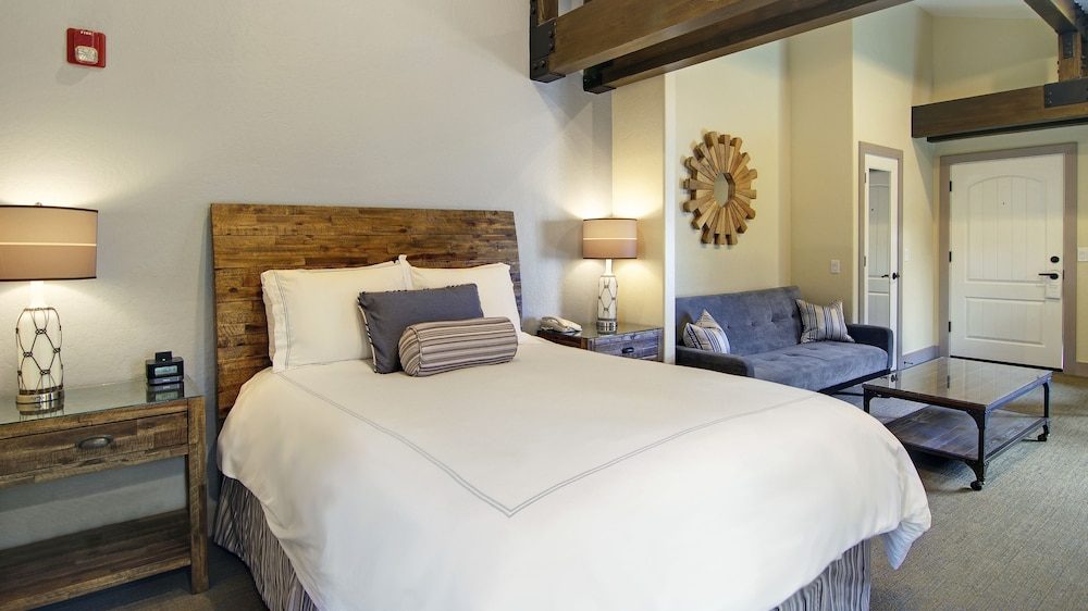 A bedroom with a large wooden headboard bed dressed in white linens and decorative pillows. On either side of the bed are wooden nightstands with modern lamps. A gray upholstered sofa with striped pillows sits against the wall near a wooden coffee table. A decorative wooden sunburst mirror hangs above the sofa. The room has beige walls, carpeted flooring, and exposed wooden beams on the ceiling. A white door is visible in the background.